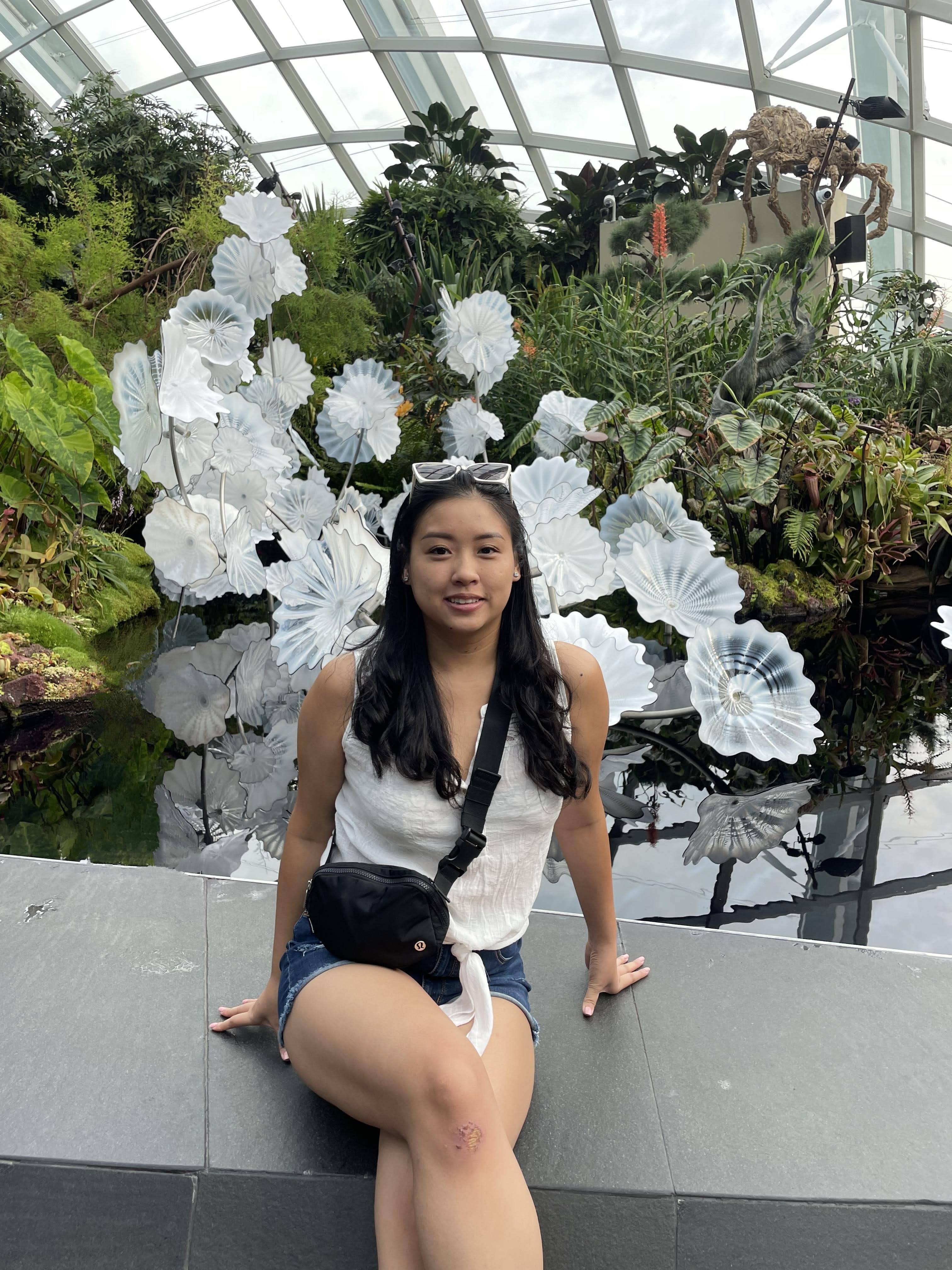 Asian woman with black hair in a white top, denim shorts, and a black belt bag, sitting in front of a fountain.